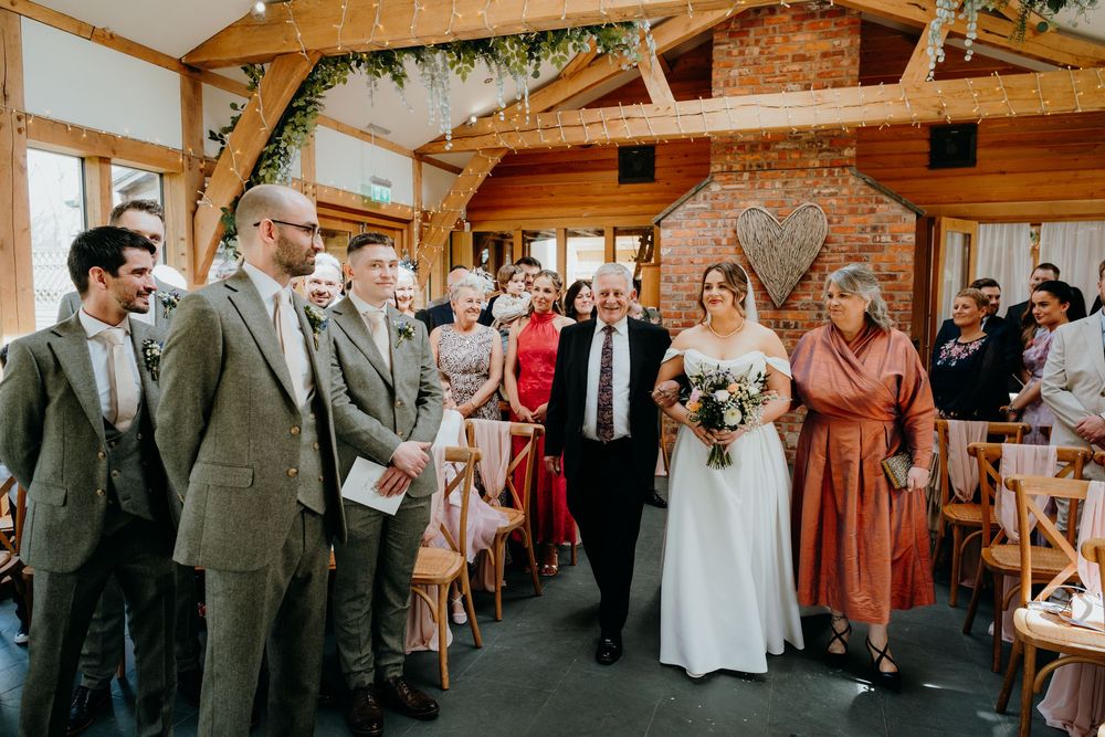 Bride walks down the aisle in a rustic barn wedding venue decorated with fairy lights and greenery.