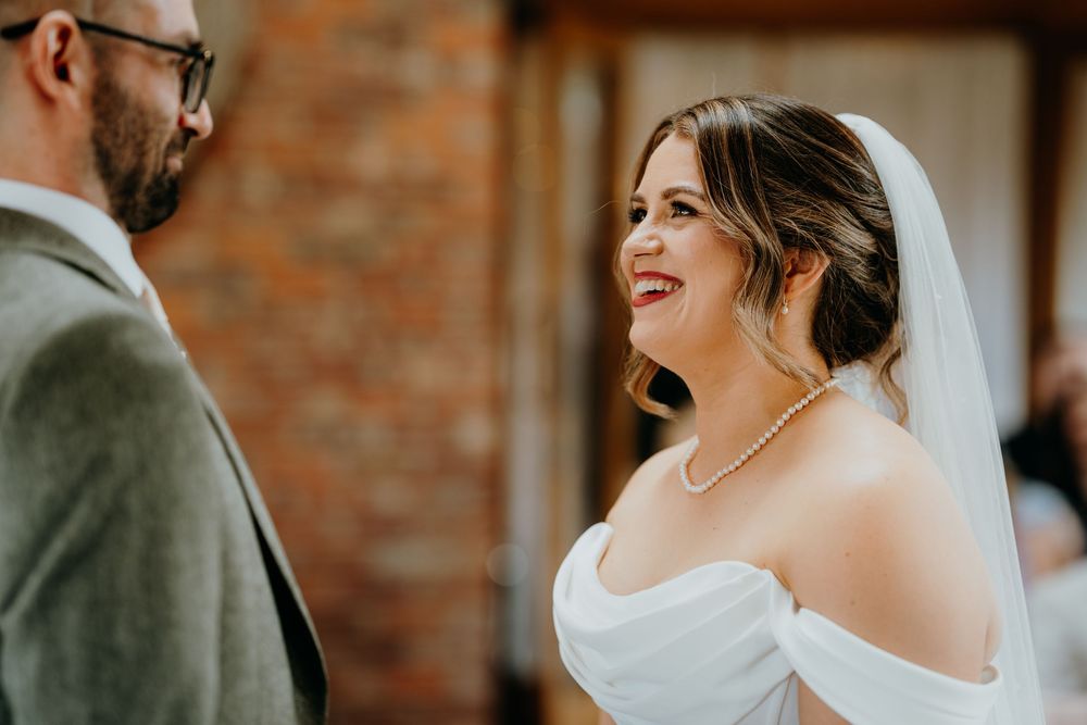 Radiant bride in white off-shoulder dress and veil smiles joyfully at groom during wedding ceremony.