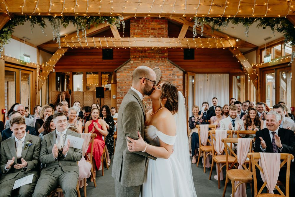 Newlyweds share their first kiss at the altar in a warmly lit barn venue with fairy lights.