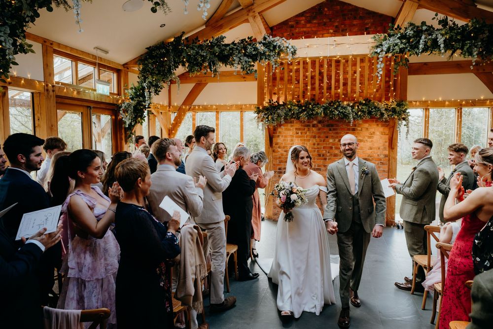 Newlyweds walk back down the aisle as guests applaud in a beautifully decorated barn wedding venue.