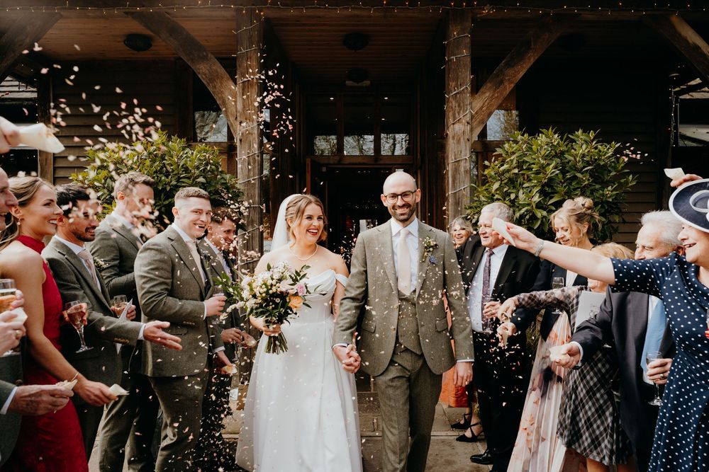 Bride and groom walk through a shower of confetti thrown by smiling wedding guests outdoors.