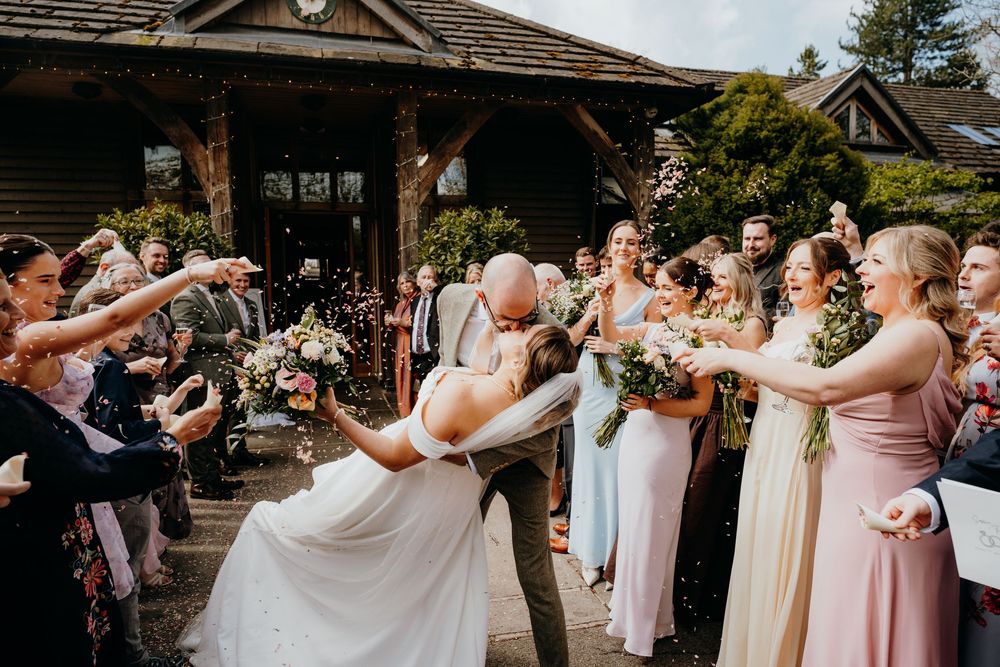 Groom dips bride for a romantic kiss as bridesmaids in pastel dresses toss confetti outdoors.