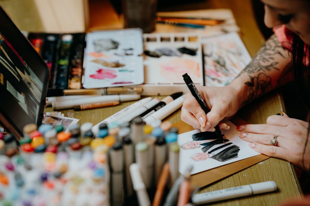 Tattooed artist draws with markers surrounded by colorful art supplies and watercolor sketches.