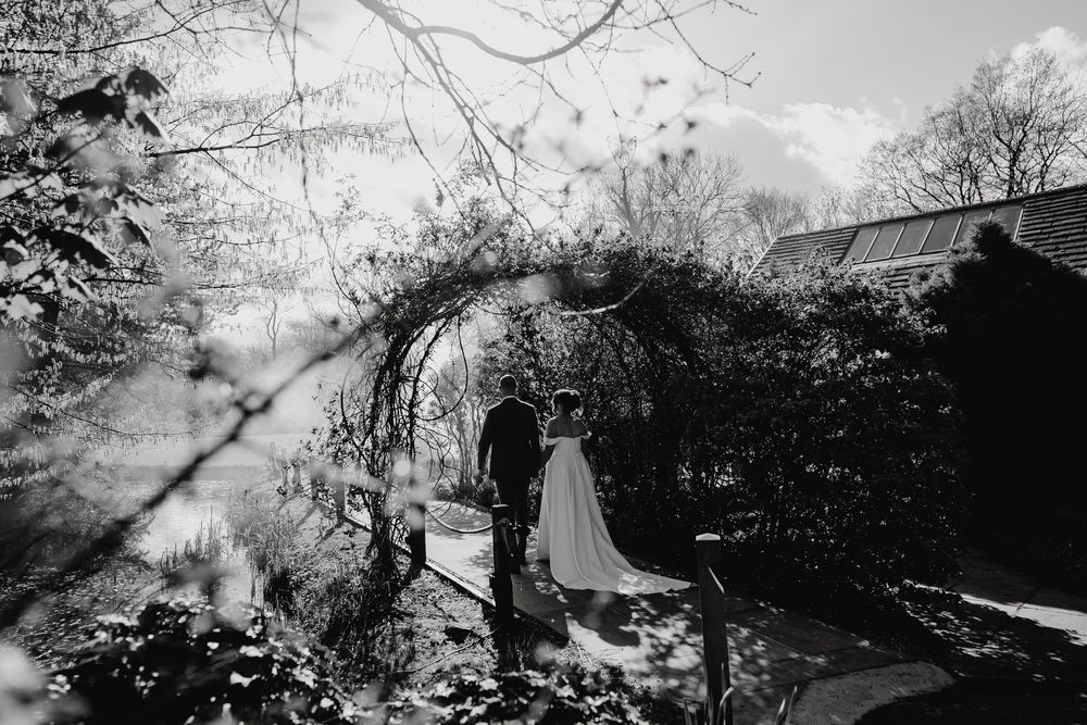 Black and white photo of bride and groom standing under a garden arch surrounded by lush foliage.
