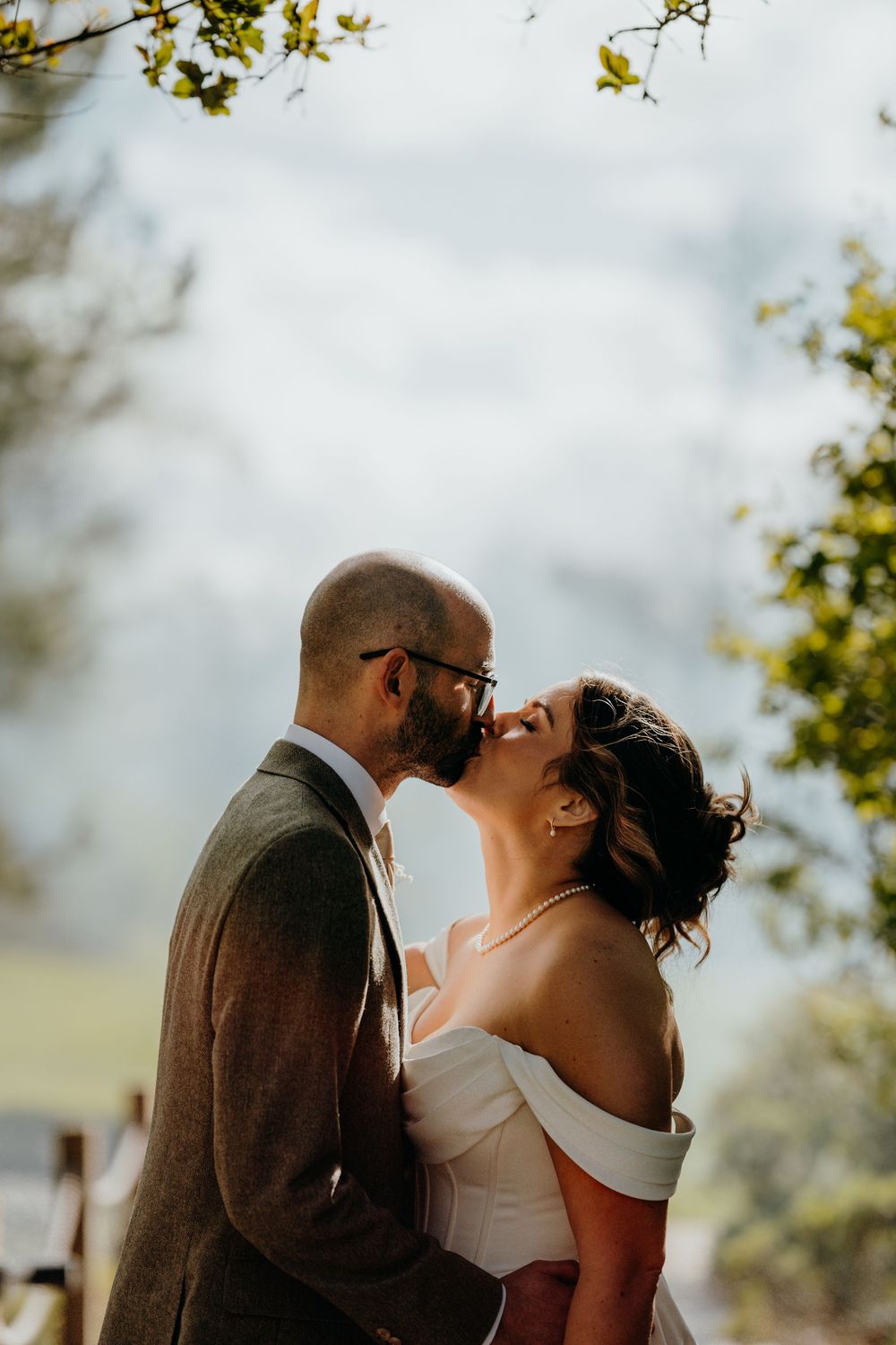 Bride and groom sharing a romantic kiss outdoors under green trees on their wedding day.