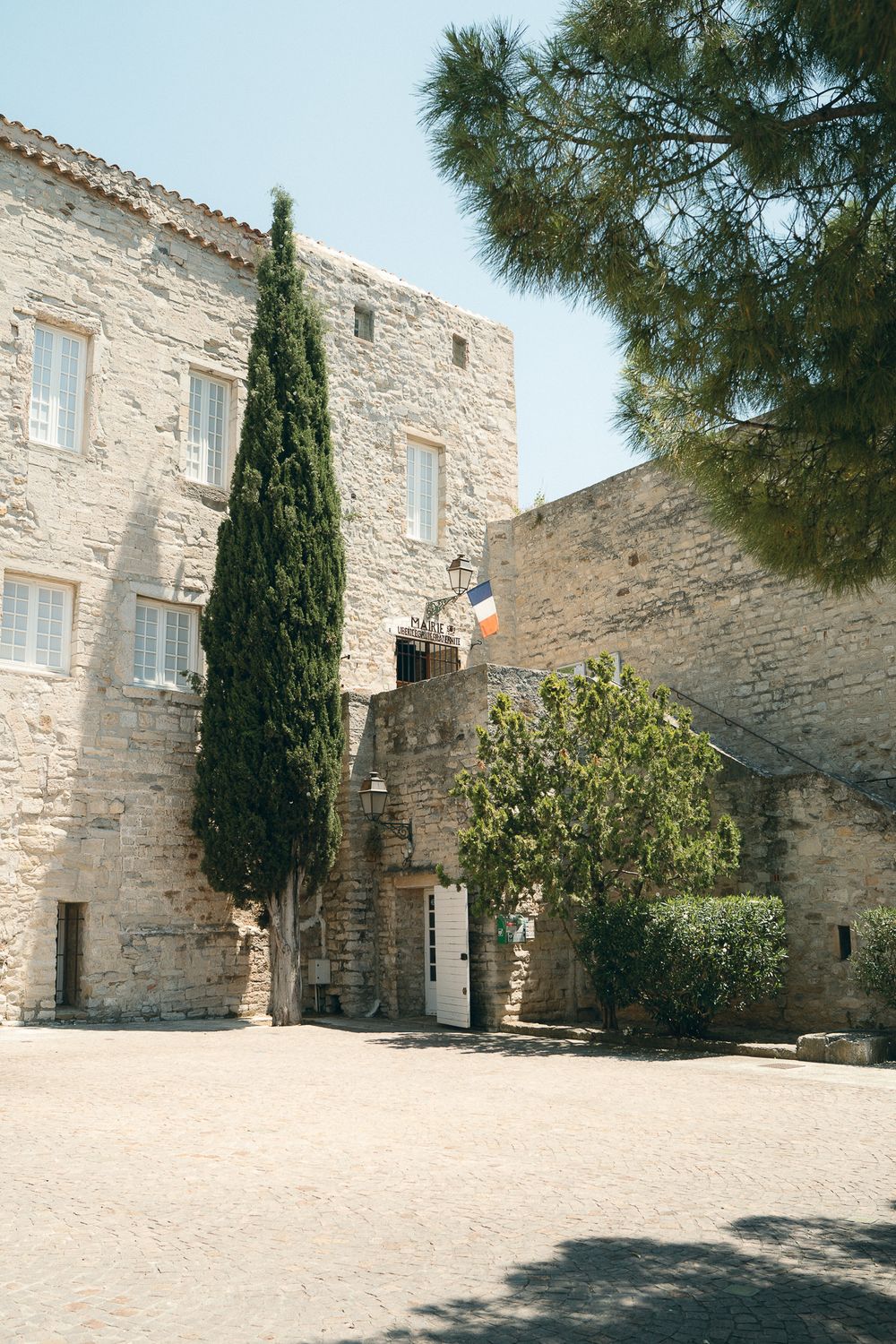 Historic stone building in southern France with cypress tree and French flag under clear blue sky.
