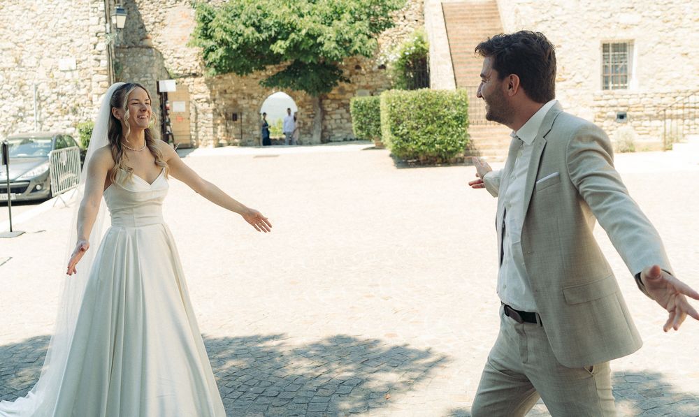 Bride in white gown and groom in tan suit share a joyful moment outdoors at a sunlit European courtyard.