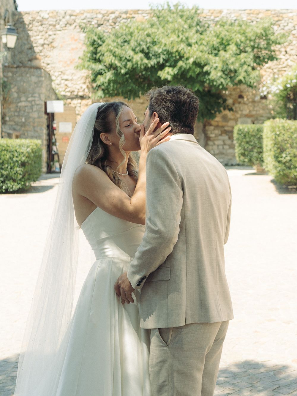 Bride and groom sharing a romantic kiss outdoors, bride in white gown with veil, groom in beige suit, stone building backdrop.