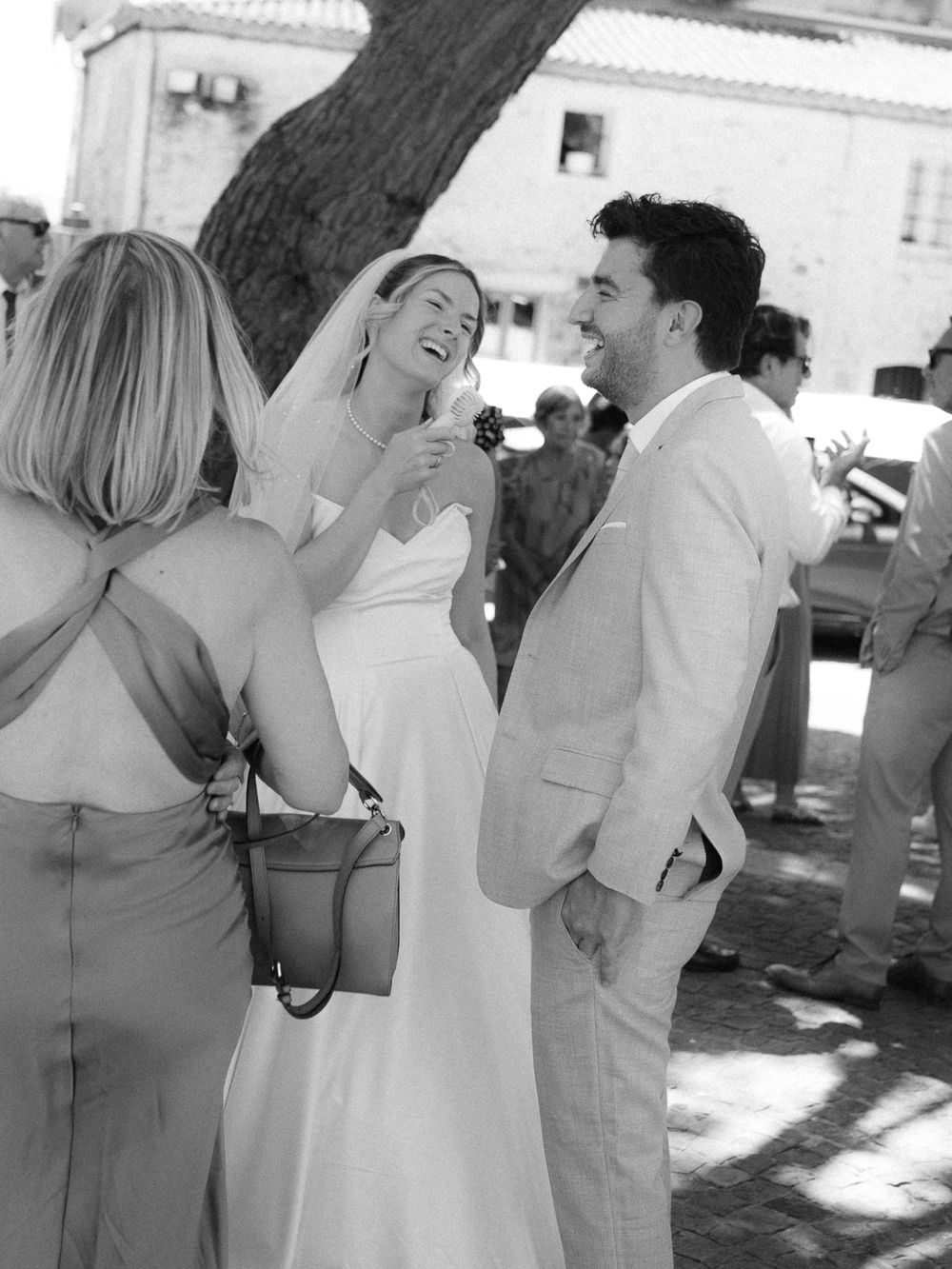 Black and white photo of a laughing bride in veil and groom in light suit chatting with guests at an outdoor wedding.