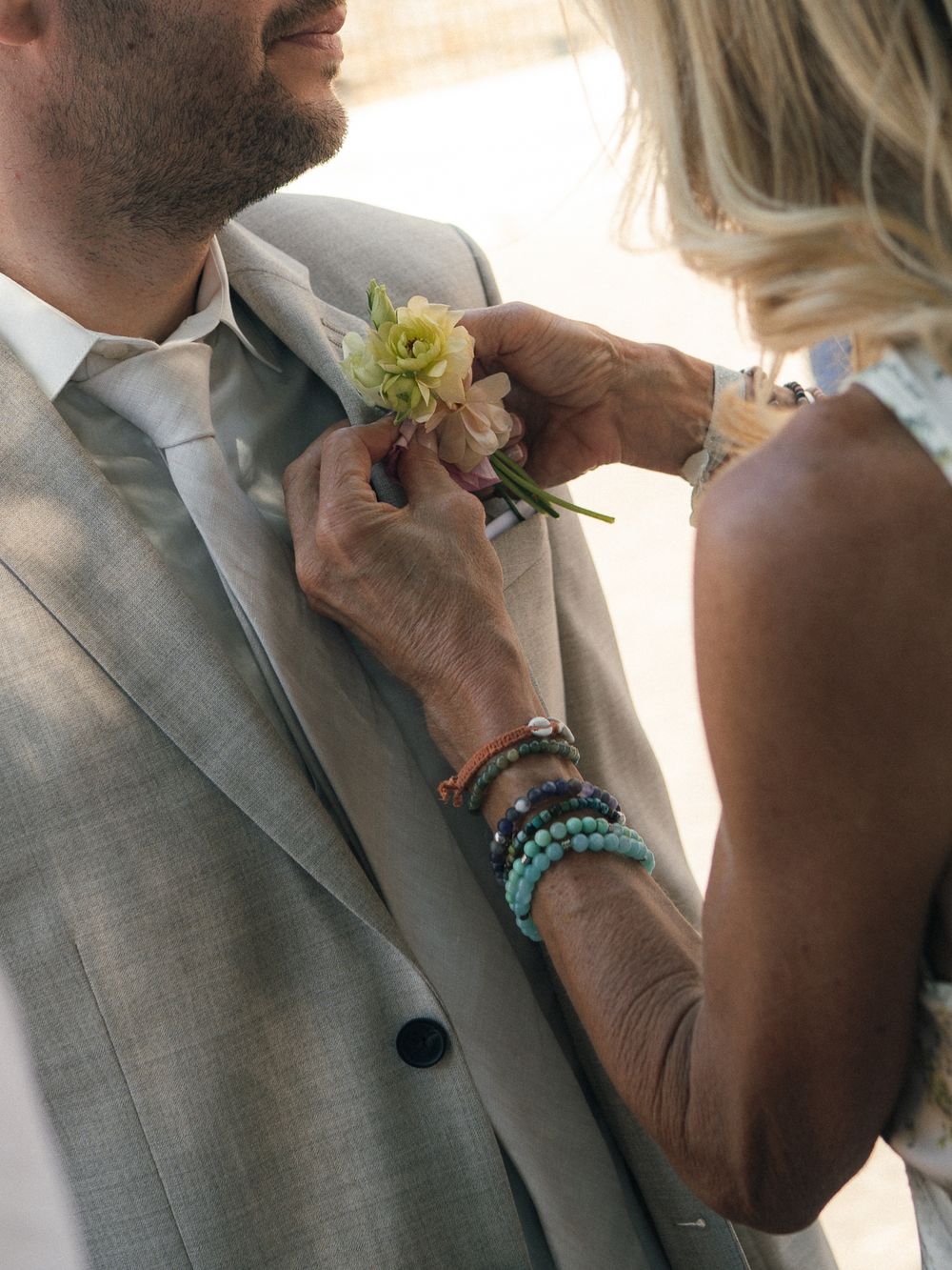 A woman with turquoise bracelets pins a floral boutonniere on a man in a gray suit and tie.