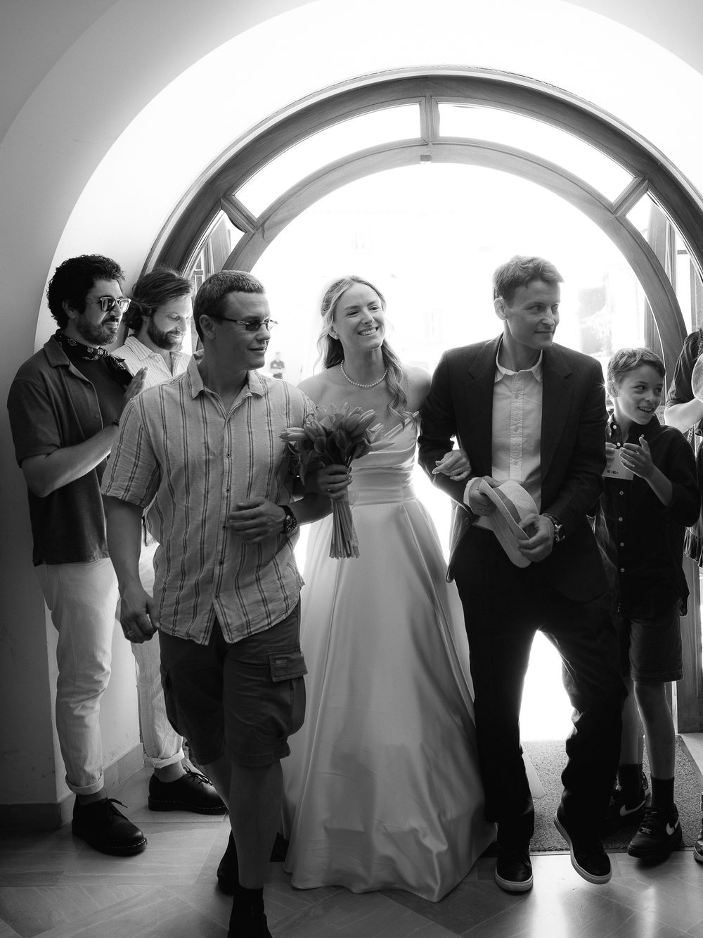 Black and white wedding photo of bride in white gown with bouquet walking through arch with groom and guests.