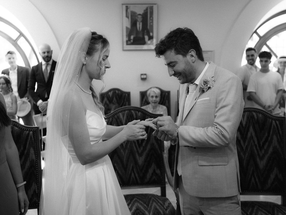Black and white photo of a bride and groom exchanging rings during a wedding ceremony indoors.