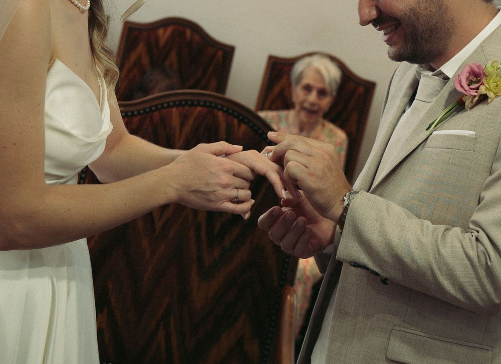 Bride placing wedding ring on groom's finger during intimate ceremony with elderly guest watching in background.