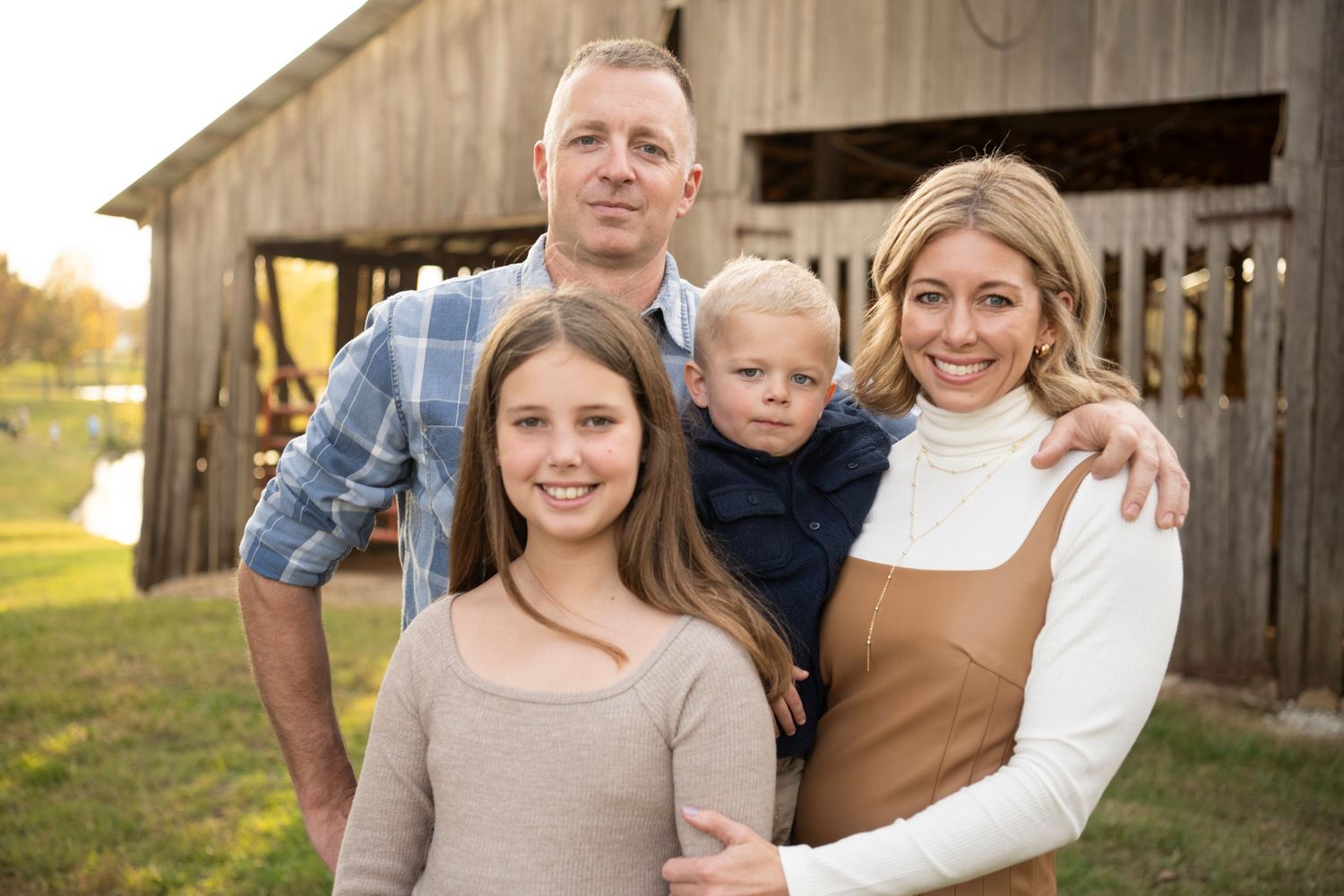 Family of four enjoying golden hour on a farm with a silo, children playing in green grass at sunset.