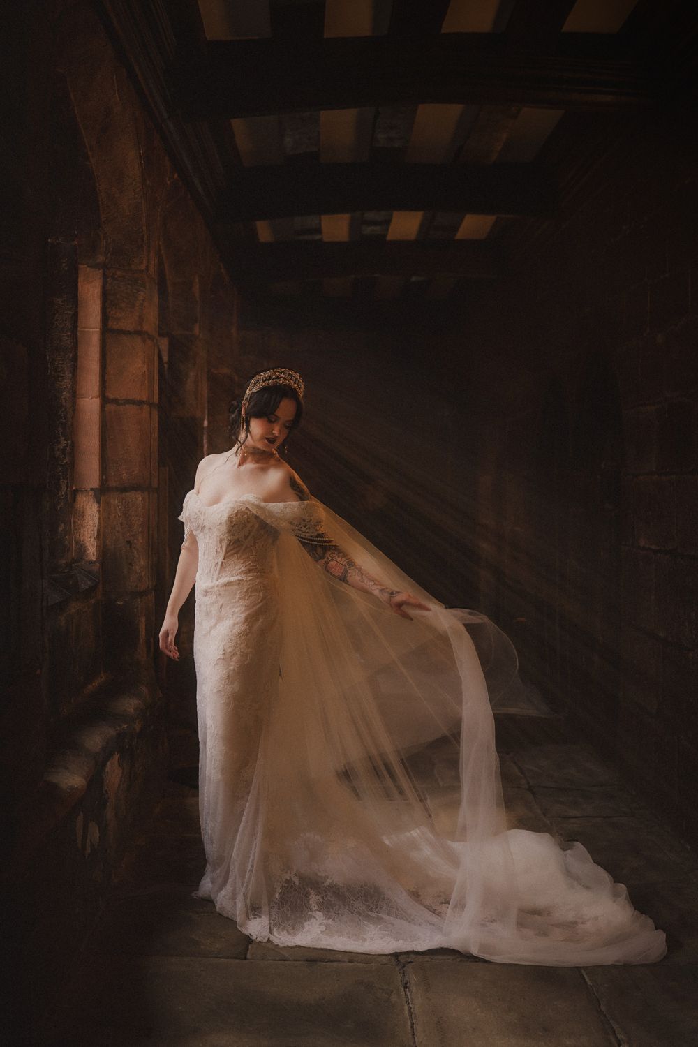Bride in flowing white tulle gown with crown stands in dramatic stone hallway with warm light rays.