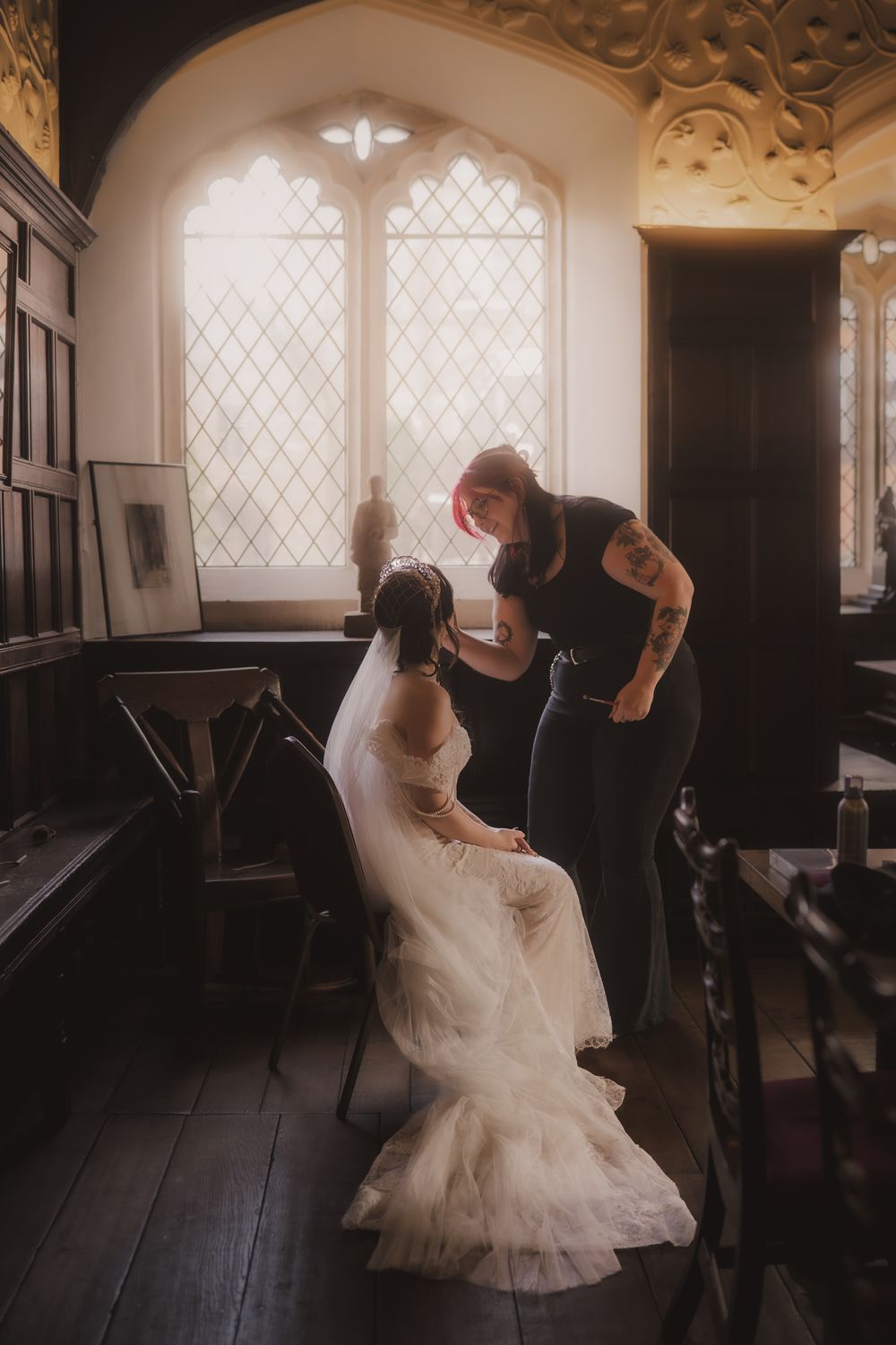 Bride in white gown seated by gothic window as tattooed stylist adjusts veil in elegant wood-paneled room.