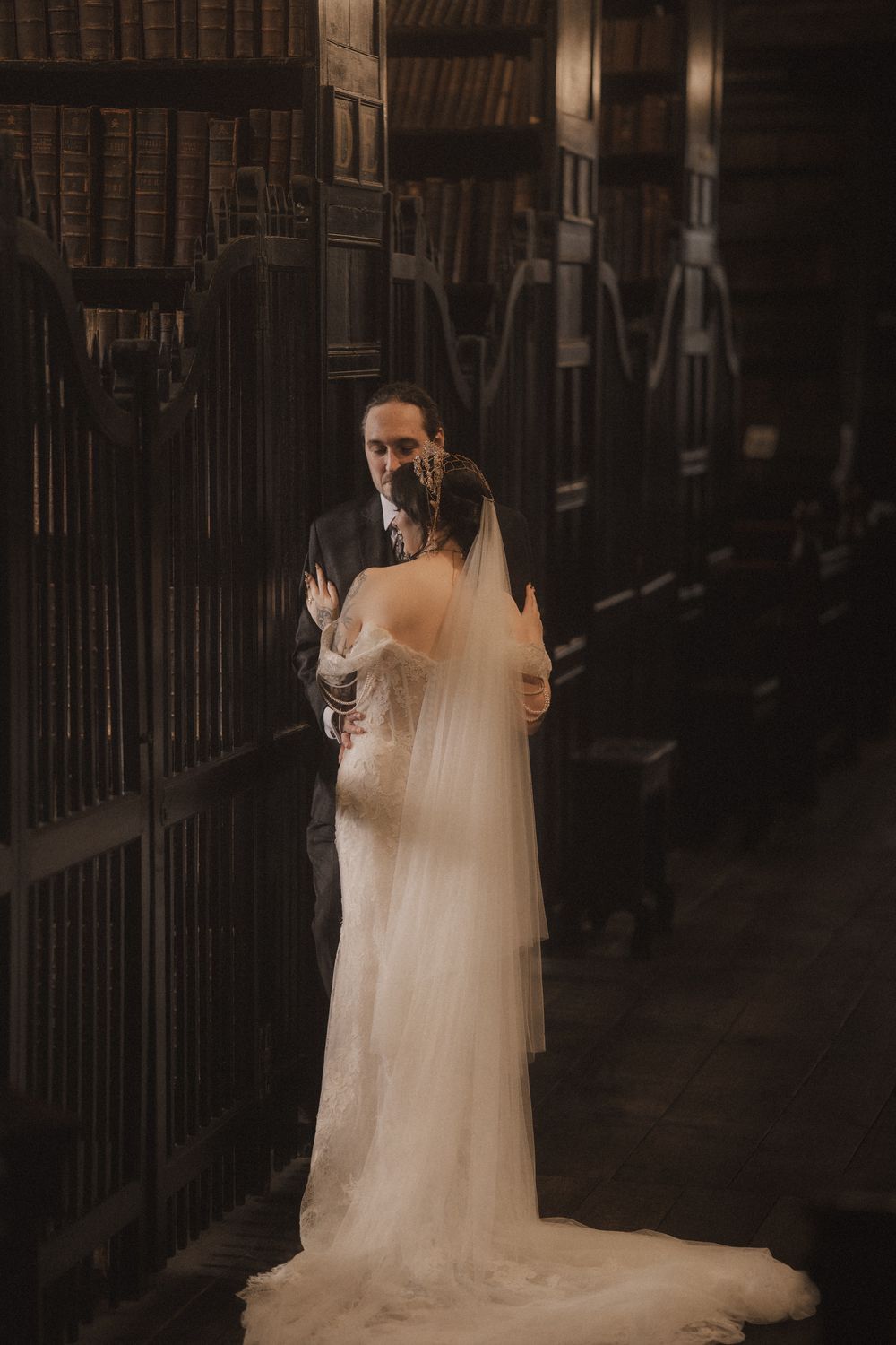 A bride in a flowing white gown and veil embraces her groom in a grand, dark wood library aisle.