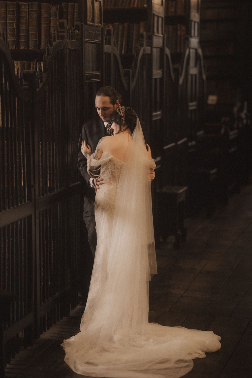 Bride and groom share a romantic embrace in a dark, elegant library aisle, bride wearing a flowing white gown with veil.