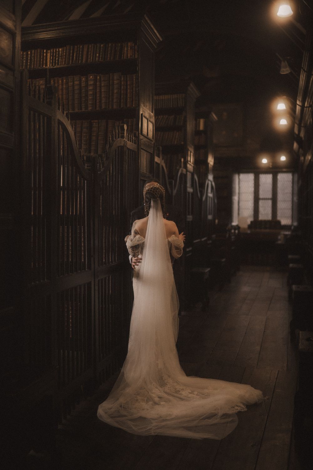 Bride in white gown with long veil stands in a dark, vintage library with tall wooden bookshelves and warm lighting.