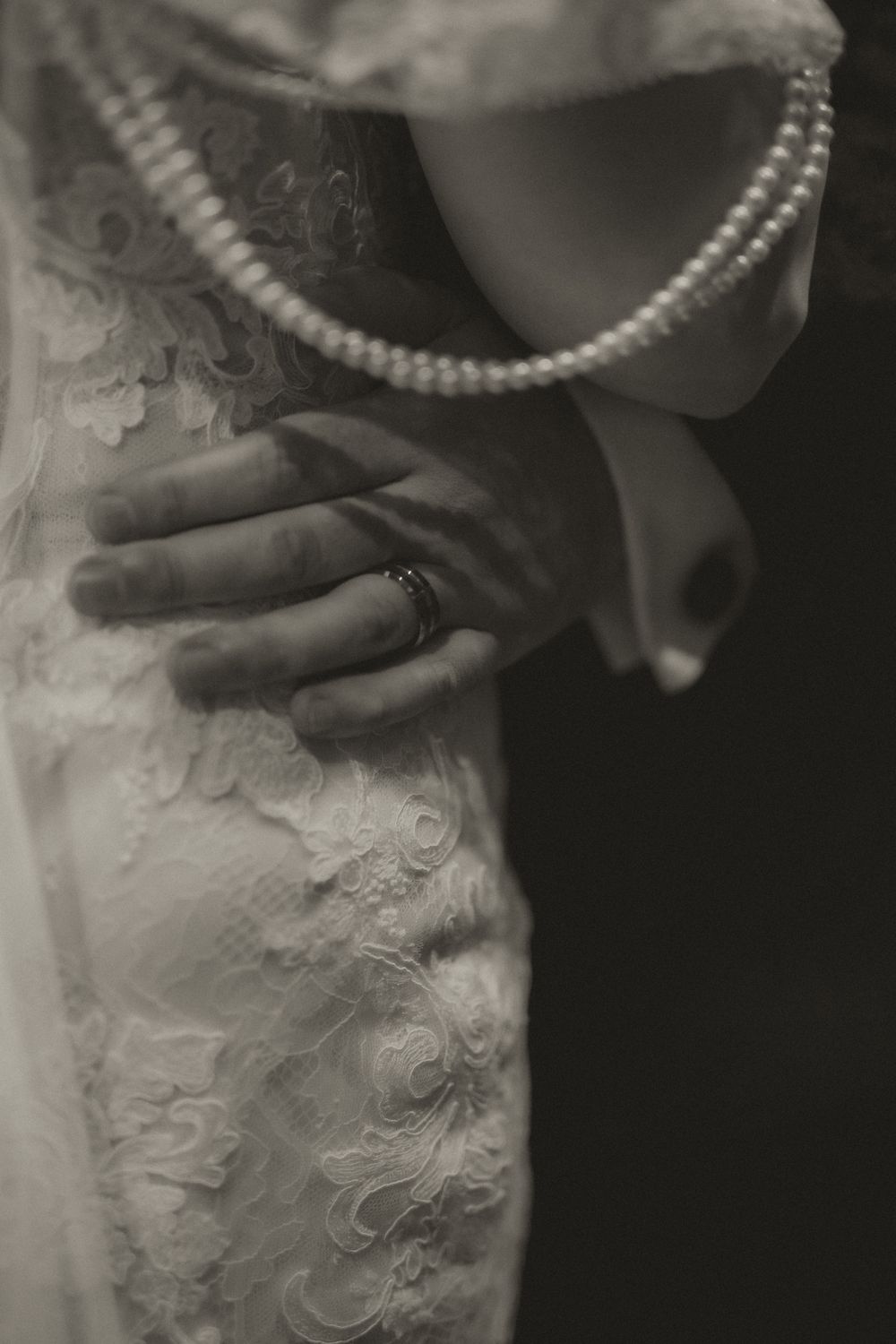 Black and white photo of a hand with a ring resting on a lace wedding dress beside a pearl necklace.
