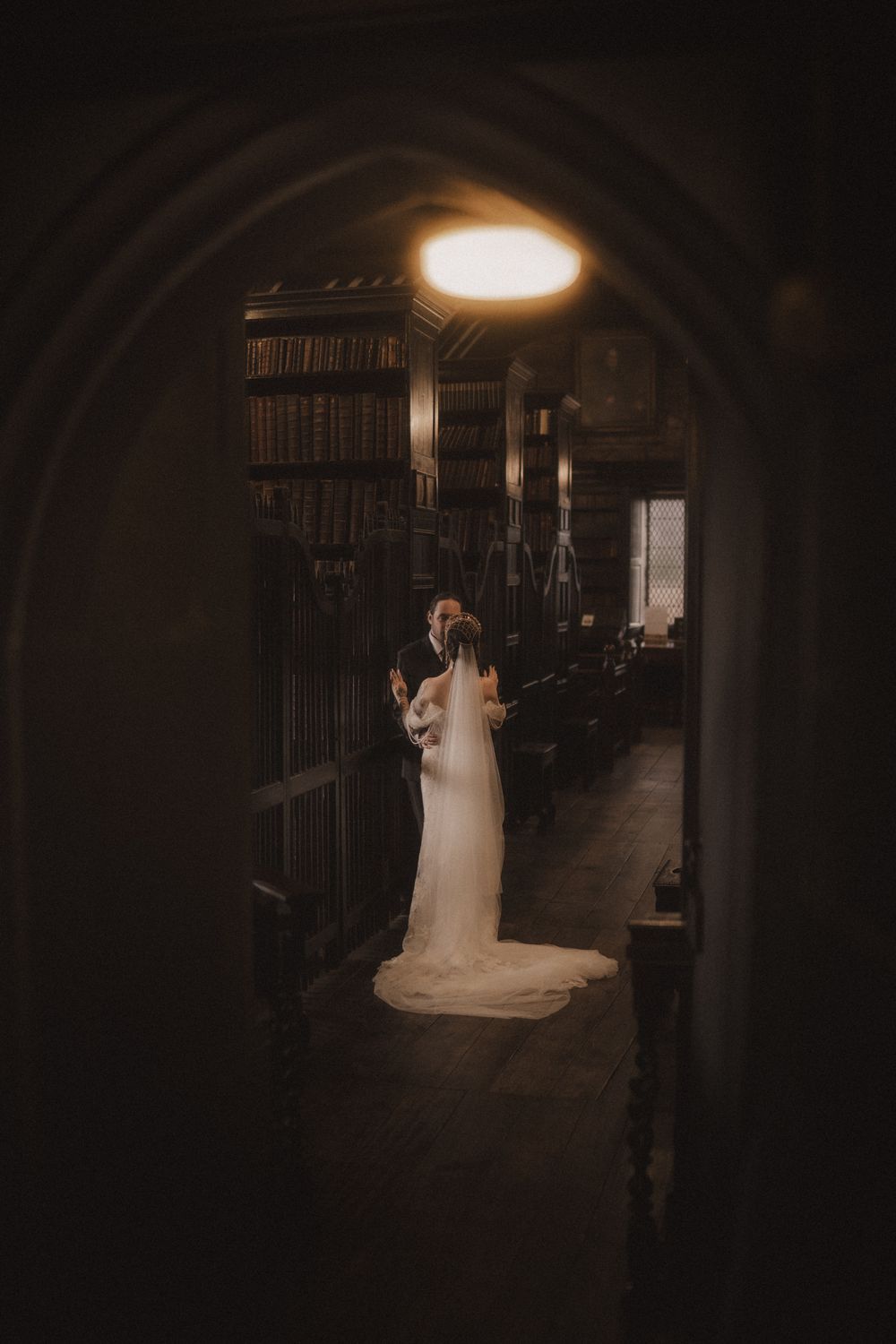 A bride and groom embrace in a grand library, framed by a dark archway with tall bookshelves and warm lighting.