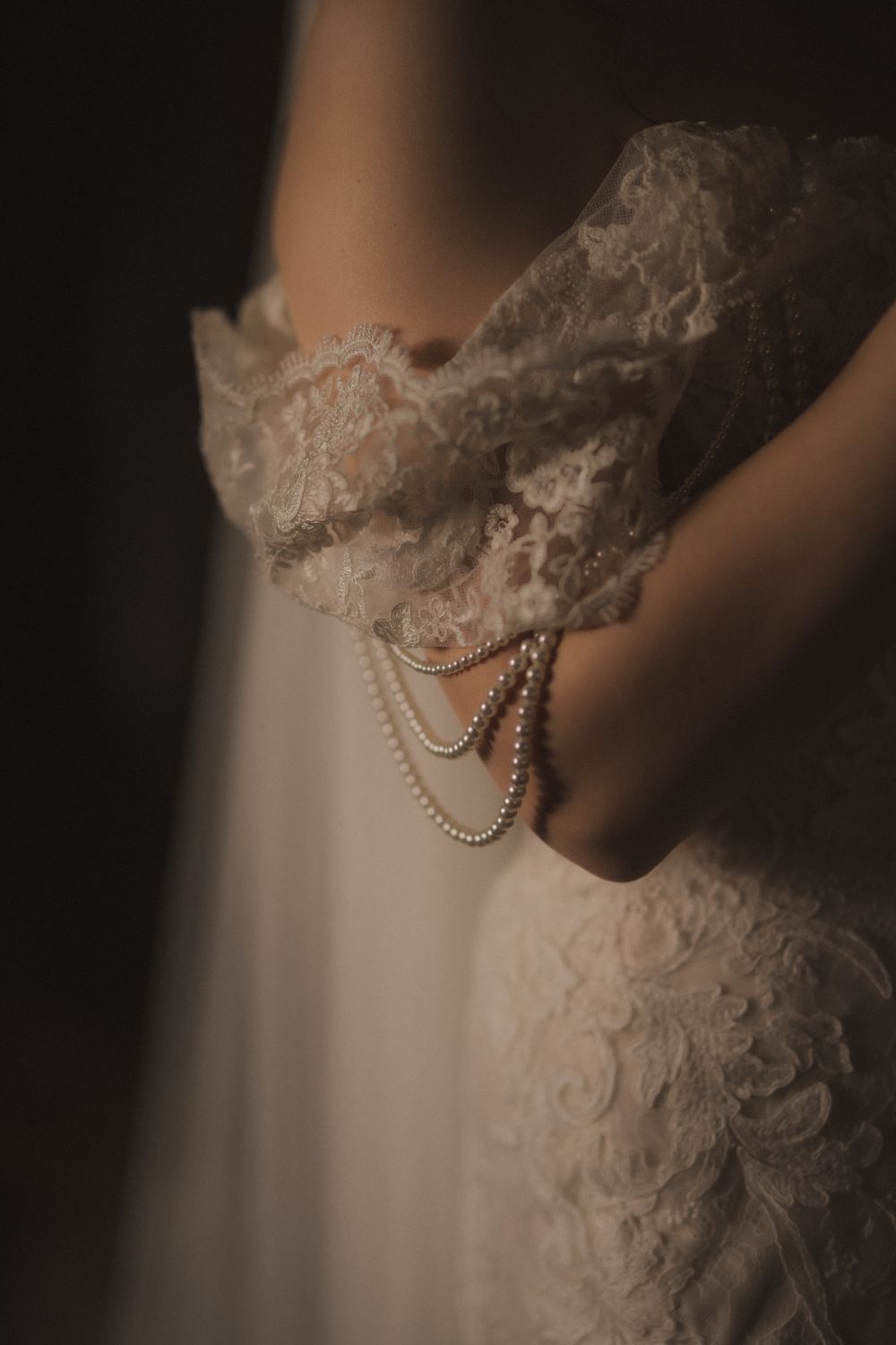 Bride in lace off-shoulder wedding gown holding pearl jewelry, moody dark background.