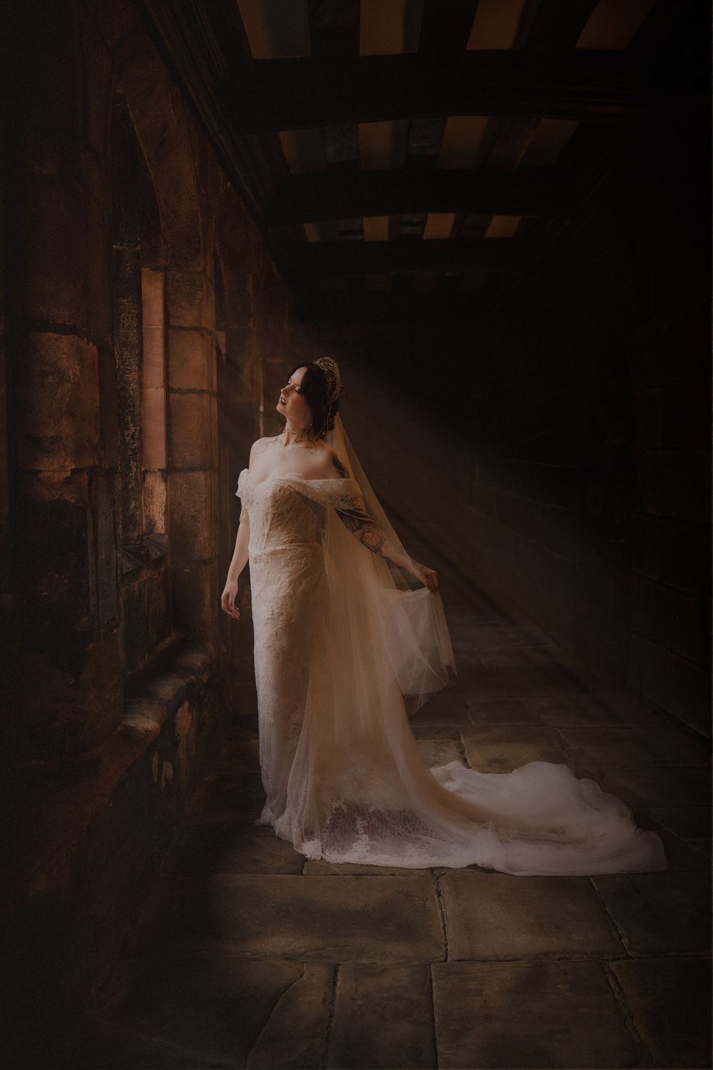 Bride in elegant white lace gown stands in dramatic dark stone corridor with light beam, creating ethereal wedding portrait.