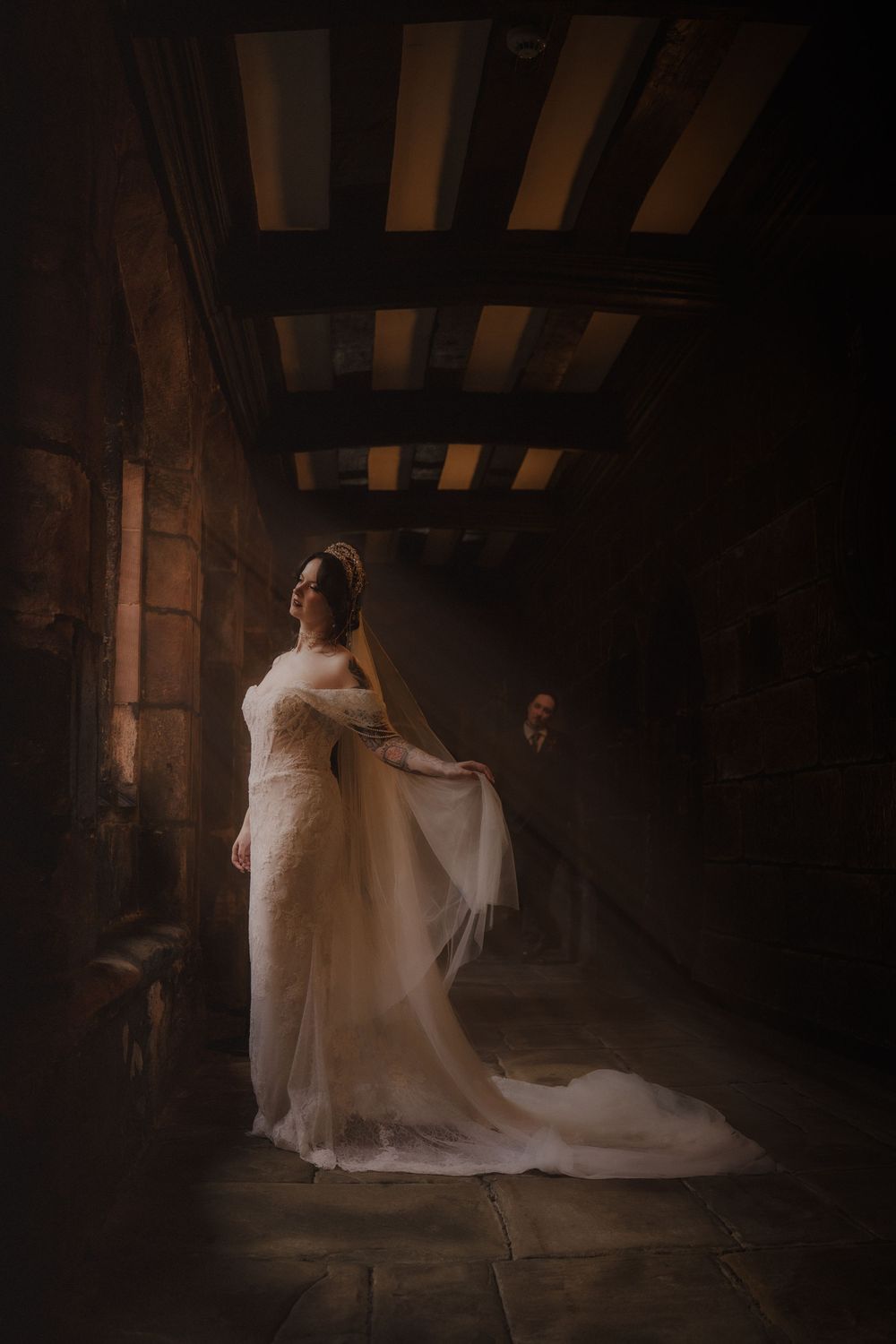 Bride in elegant white lace gown with flowing veil stands in dimly lit stone castle corridor with wooden beam ceiling.