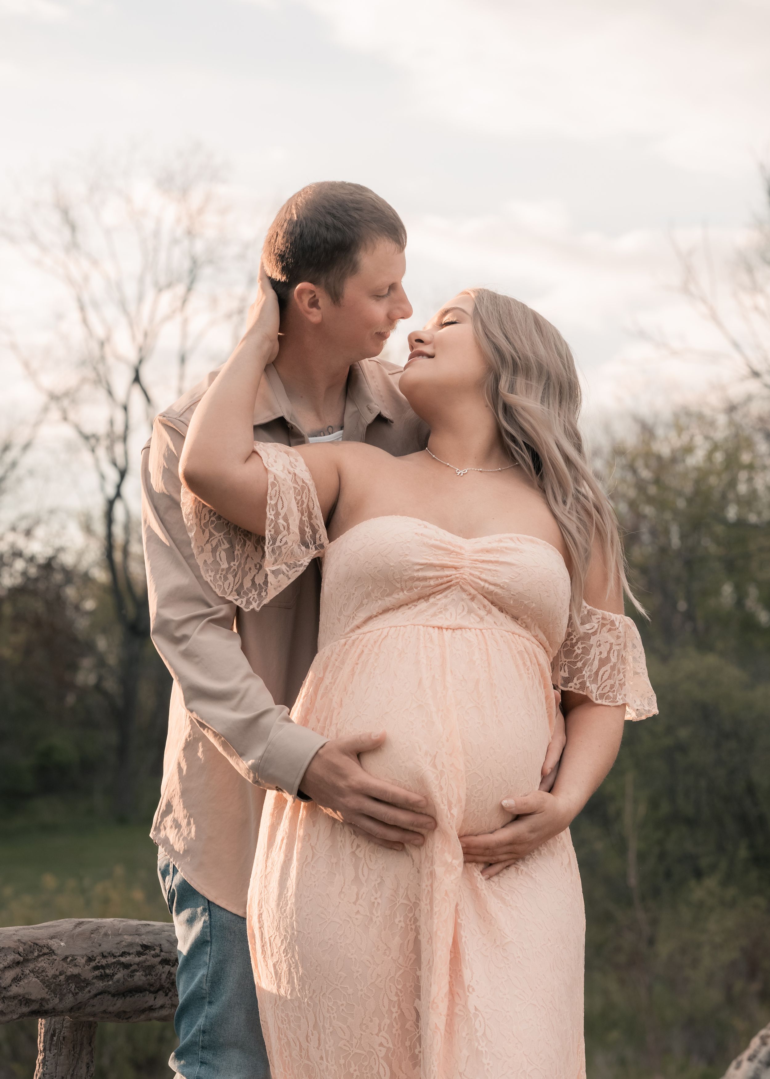 Couple sharing a tender moment outdoors, pregnant woman in pink lace maternity gown cradling baby bump.