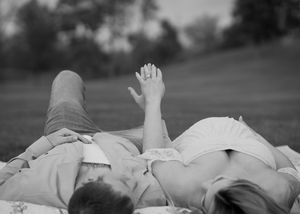 Black and white photo of a couple lying on a blanket outdoors, holding hands with an engagement ring visible.