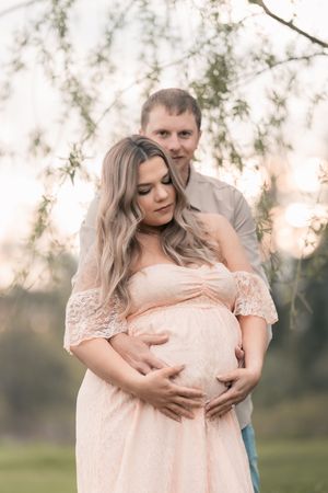 Pregnant woman in pink lace dress cradled by partner outdoors at golden hour, maternity photo shoot.