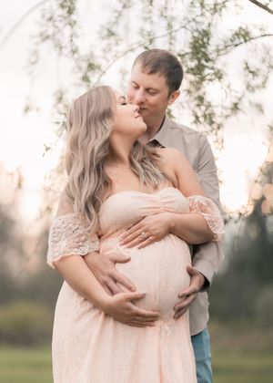 Couple sharing a tender kiss during outdoor maternity photo shoot, woman in pink lace gown cradling baby bump.