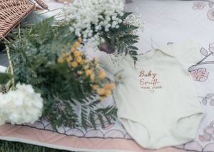 White baby onesie embroidered with Baby Smith name on a picnic blanket surrounded by yellow and white flowers.