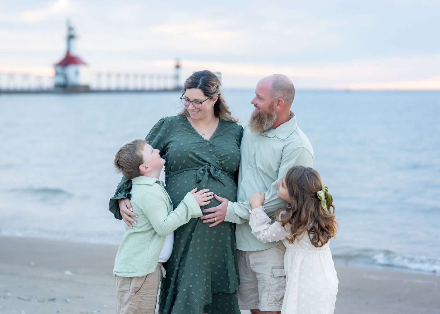 Pregnant woman in green dress with family on beach near lighthouse, husband and two children touching her baby bump.