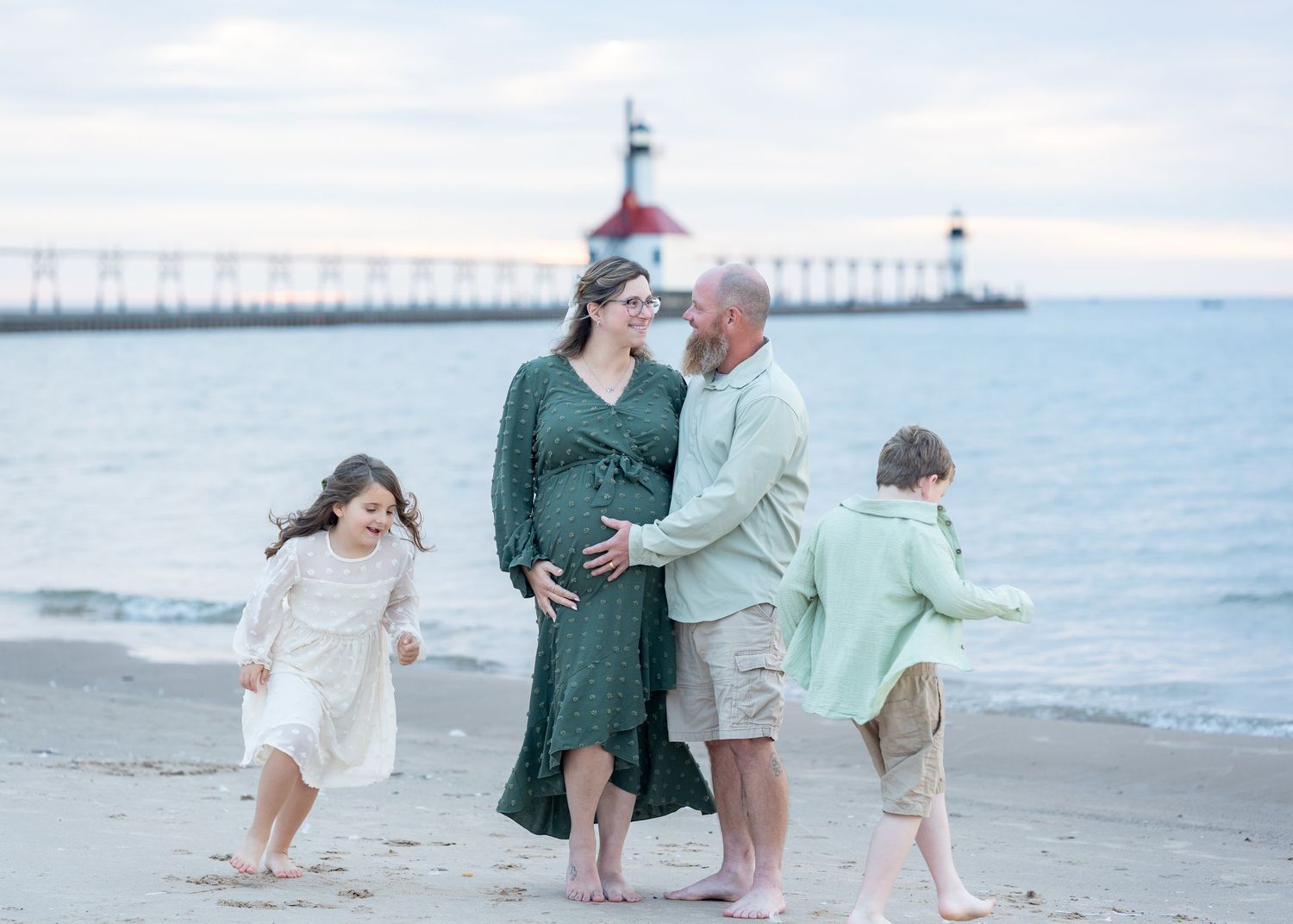 Pregnant woman and husband with two children on a beach near a lighthouse for a maternity photo session.