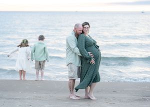 Pregnant couple embracing on beach at dusk, two children playing at water's edge, maternity photo session.