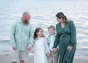 Pregnant woman in green dress with bearded husband and two children smiling on a sandy beach.