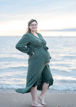 Pregnant woman in green wrap dress standing barefoot on beach at sunset, smiling with hand on baby bump.