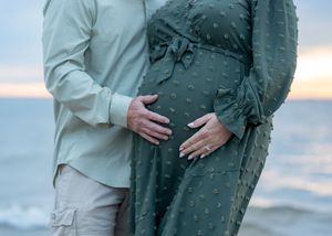Couple tenderly cradling pregnant belly at sunset beach, woman in green polka dot maternity dress.