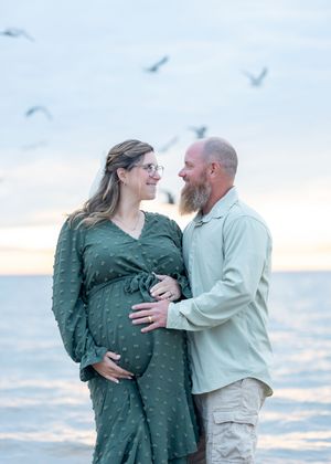 Pregnant couple embracing on beach at sunset with seagulls flying overhead, woman in green dress cradling baby bump.