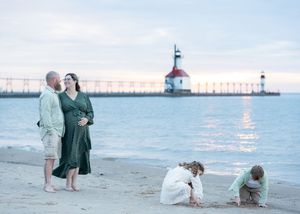 Pregnant couple embraces on beach near lighthouse while children play in sand at sunset.