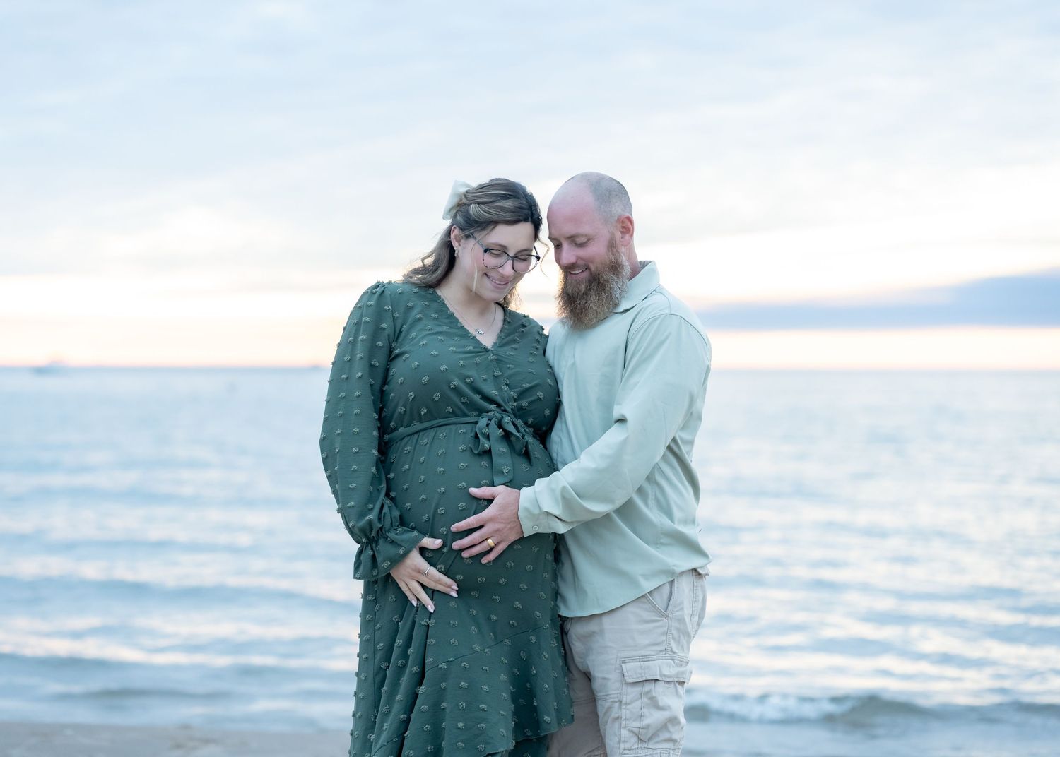 Pregnant couple embracing on a beach at sunset, woman in green dress with partner tenderly touching her baby bump.