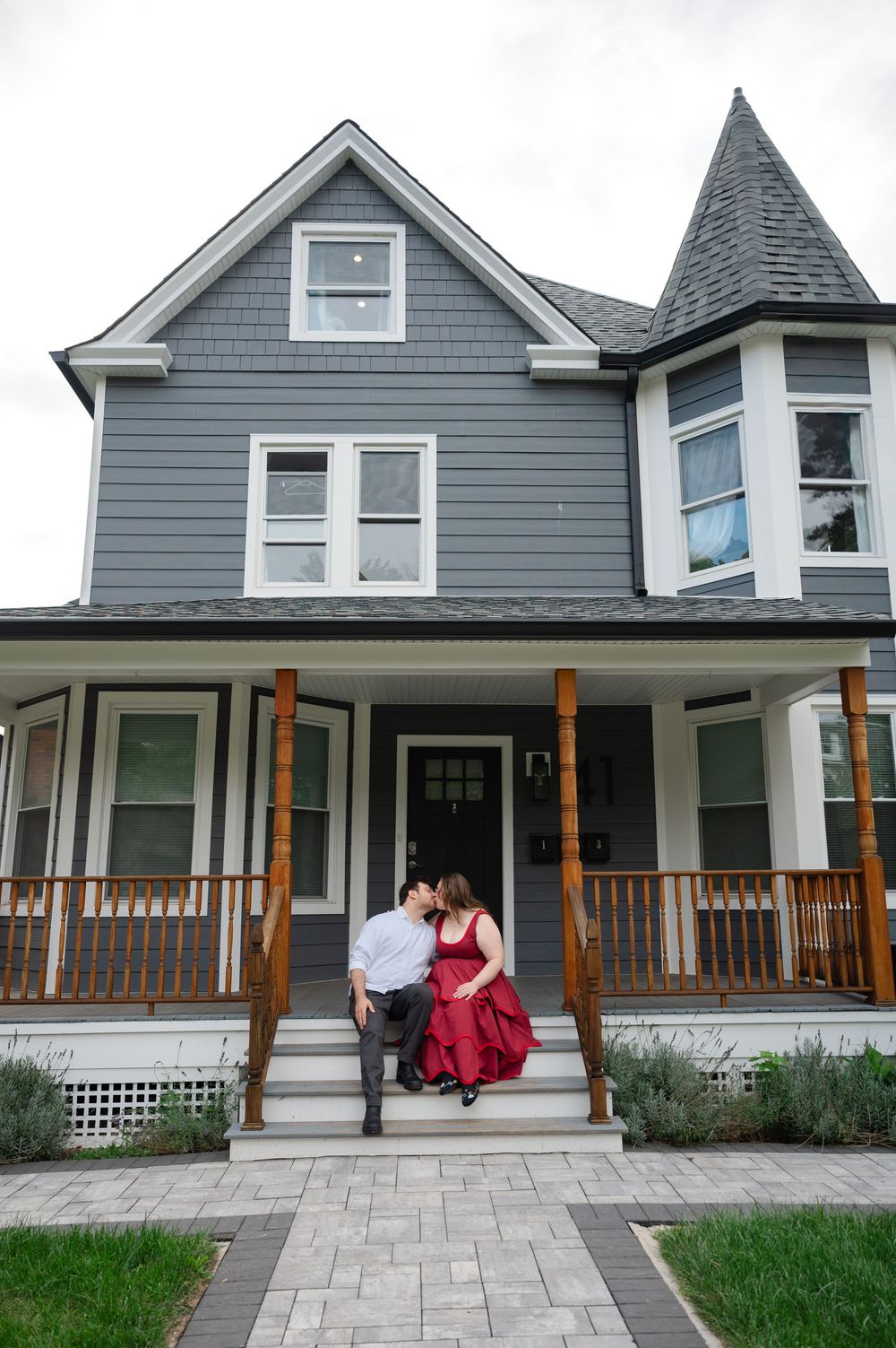 Couple sharing a romantic kiss on the porch steps of a charming gray Victorian house with white trim.
