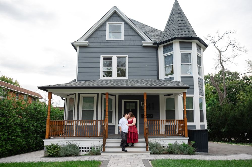 Couple embracing on the porch of a charming gray Victorian house with a turret and wooden railings.