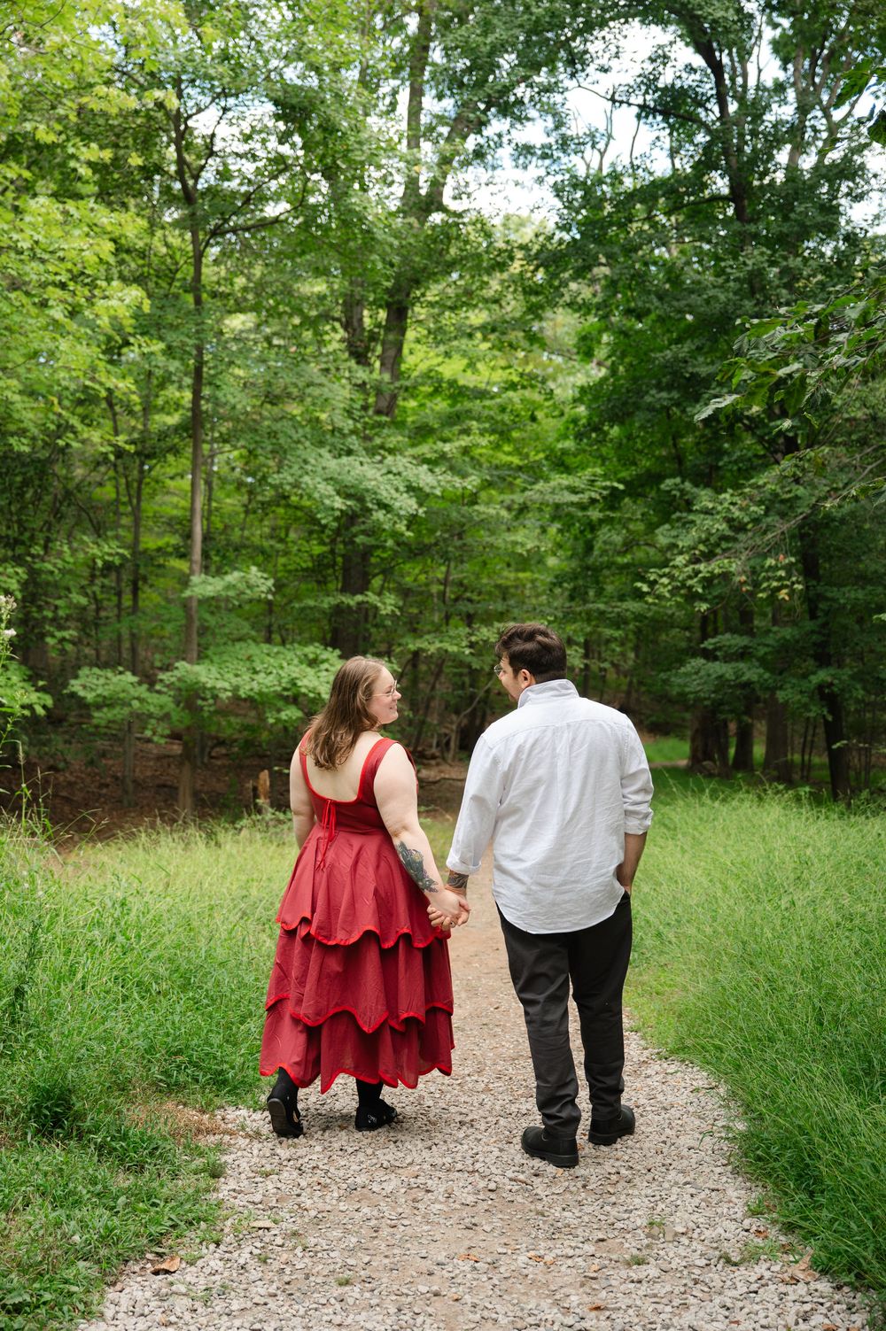 Couple holding hands walking down a wooded path, woman in red tiered dress and man in white shirt.