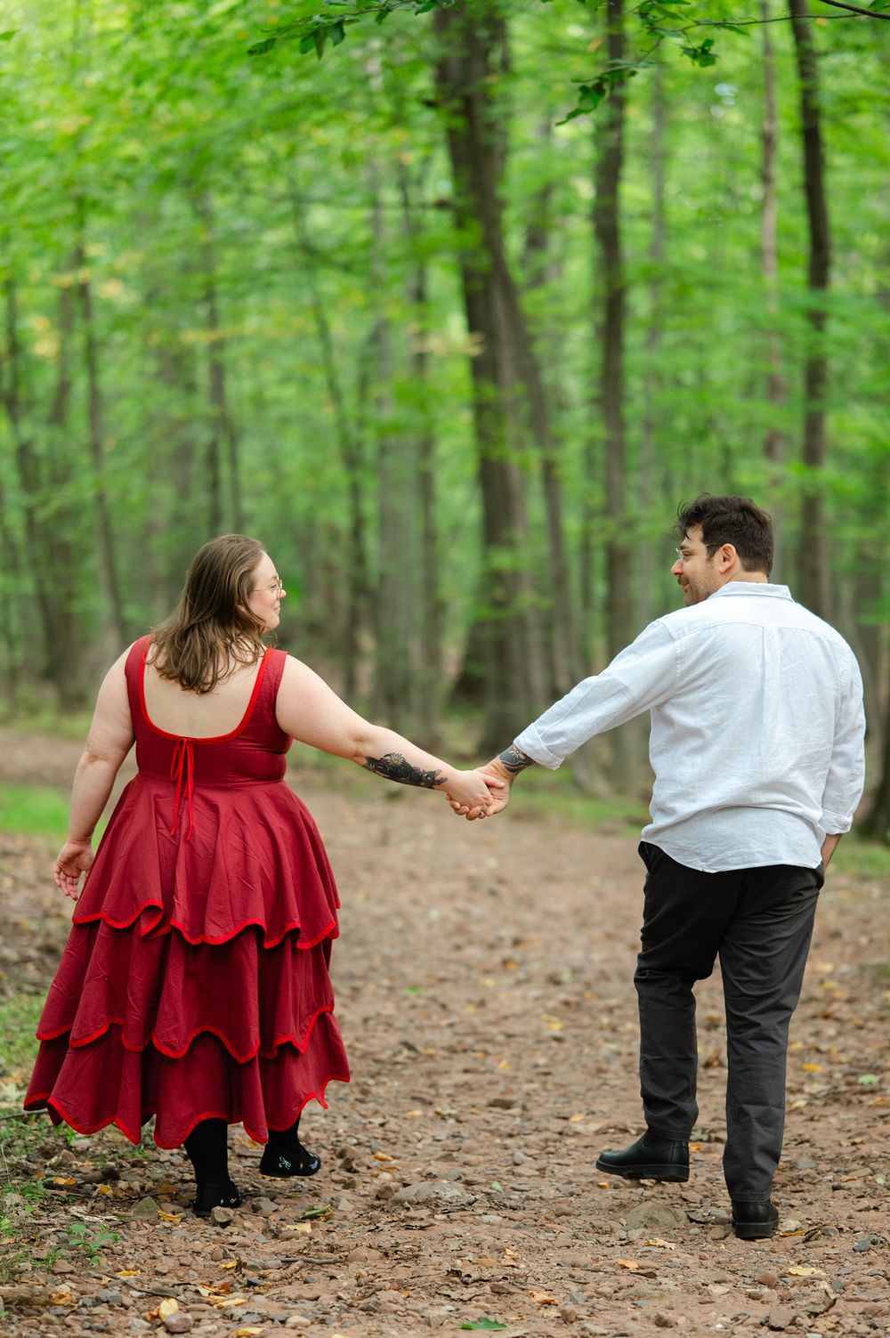 Engaged couple holding hands on a forest trail, woman in red dress looking back at her partner.