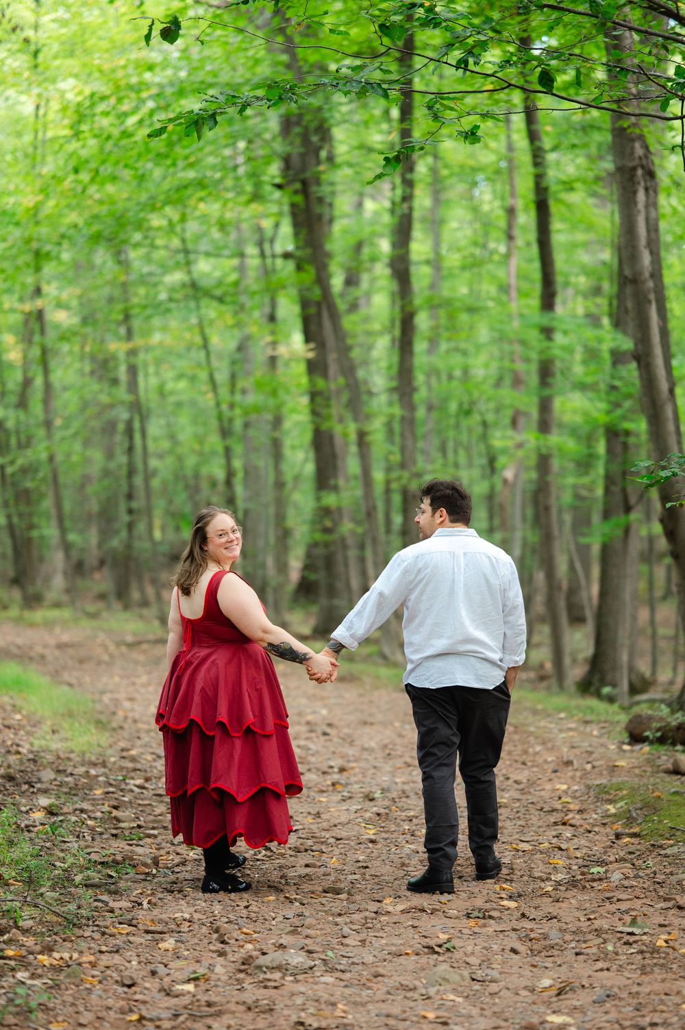 Couple holding hands walking on a forest path, woman in red tiered dress looks back smiling.