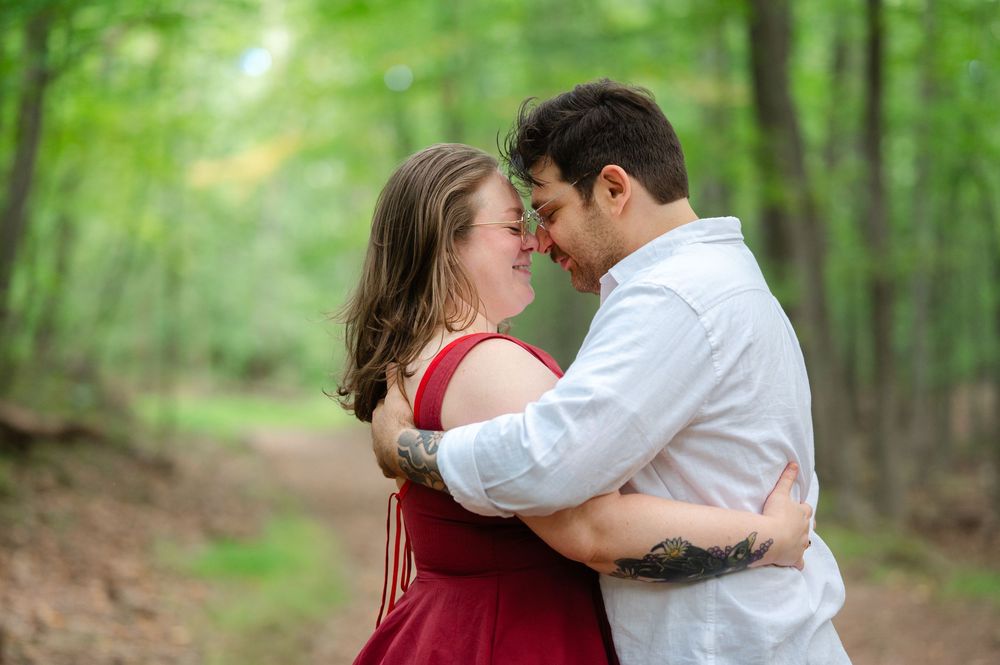 Couple embracing and touching foreheads on a forest path, woman in red dress, man in white shirt, surrounded by lush green trees.