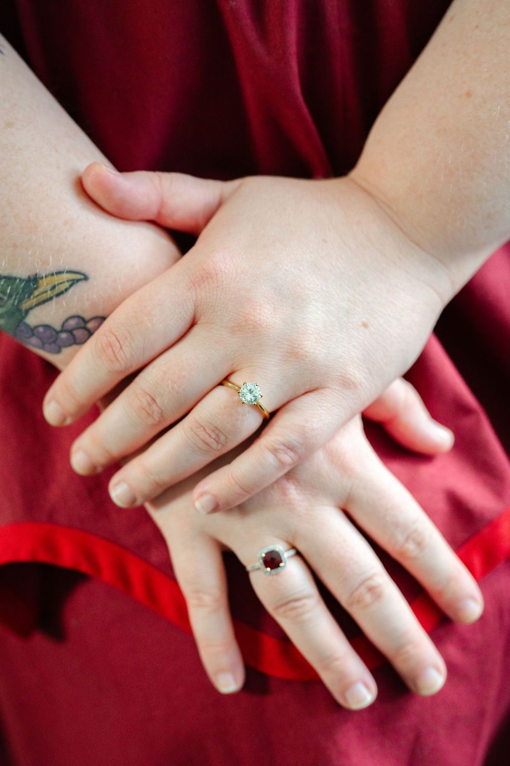 Two hands crossed wearing rings, one gold with aquamarine and one silver with garnet, against a red dress.