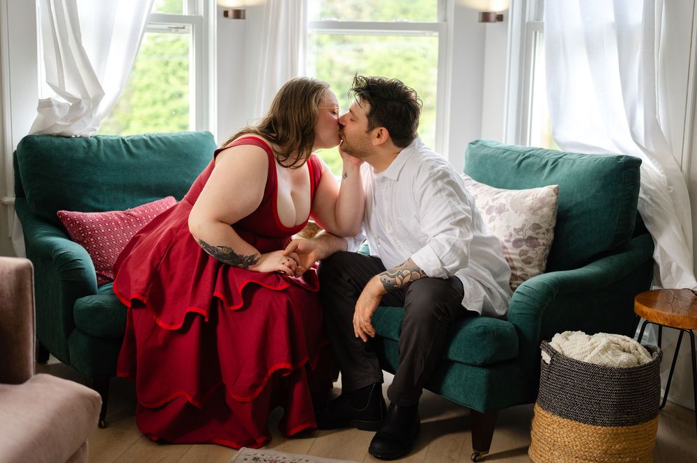 Couple sharing a romantic kiss on a teal sofa, woman in red dress, man in white shirt, bright window backdrop.